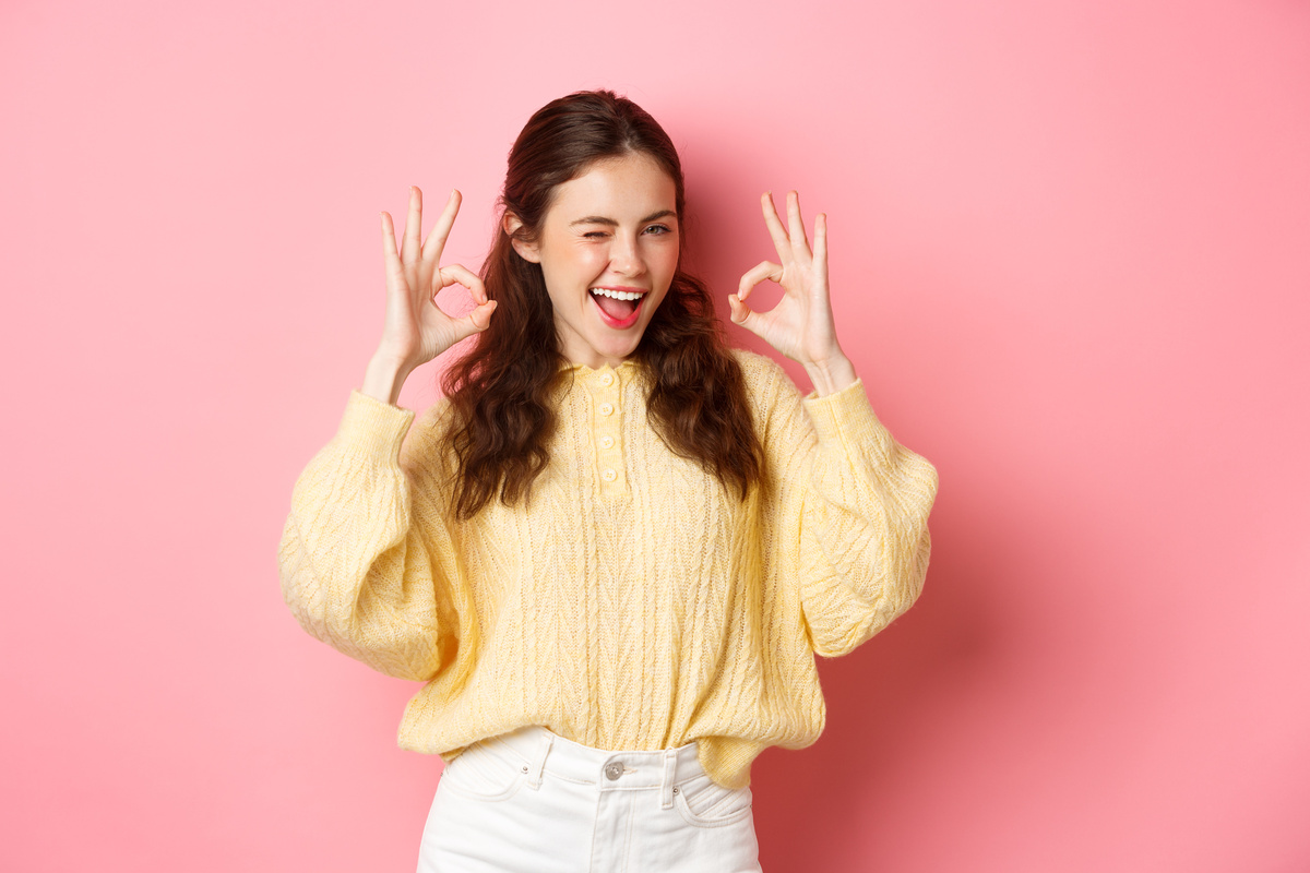 Portrait of Cheerful Pretty Girl Winking and Saying Yes, Showing Okay Sign, Alright Gesture, Praise Good Job, Well Done, Standing Pleased against Pink Background